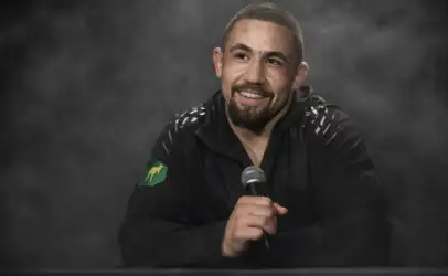 Smiling man in a black jacket holds a microphone at a desk with a smoky gray backdrop behind him.