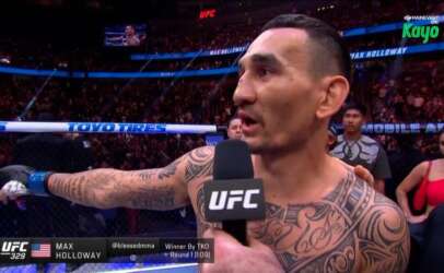 Max Holloway being interviewed in the UFC octagon after a fight, microphone labeled UFC, tattoos on chest visible, arena crowd in the background.
