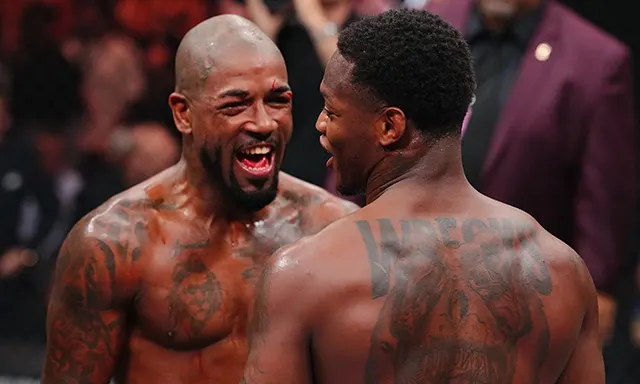 9560901c56718e49aa07722c9cd51393 - AfroCombat Two shirtless fighters with tattoos celebrate and laugh in a crowded ring, one facing the camera with a wide smile.