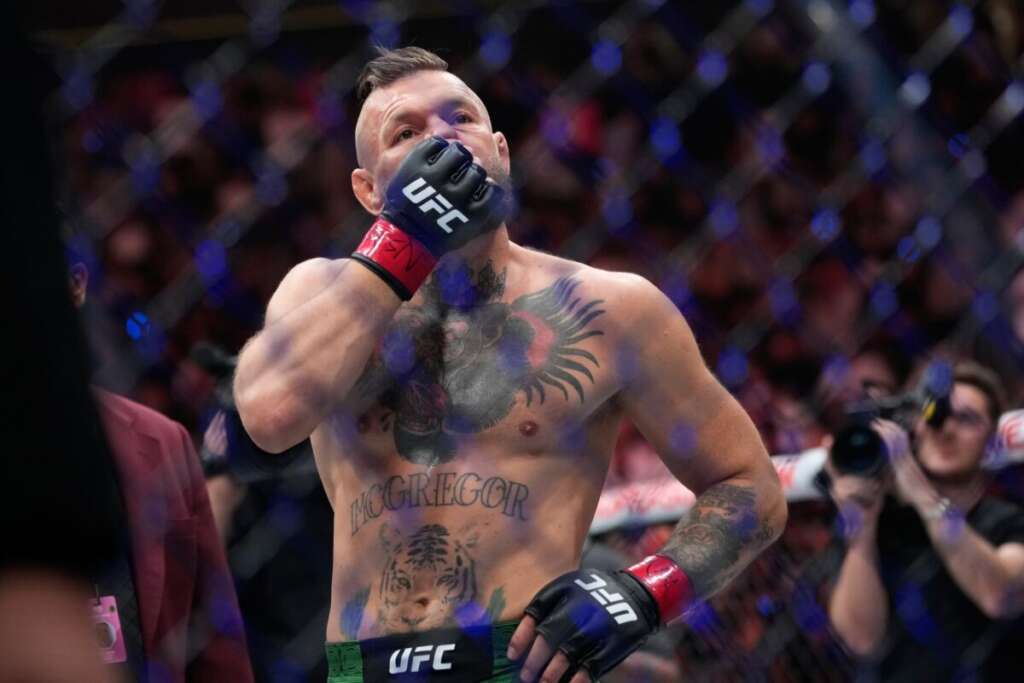 UFC fighter in gloves touches his face inside the octagon, with prominent chest tattoos and a blurred crowd in the background.