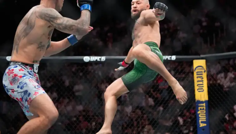 Two MMA fighters in mid-air kicks inside the UFC cage, one in green shorts attacking as the other wearing floral shorts defends with a raised glove.