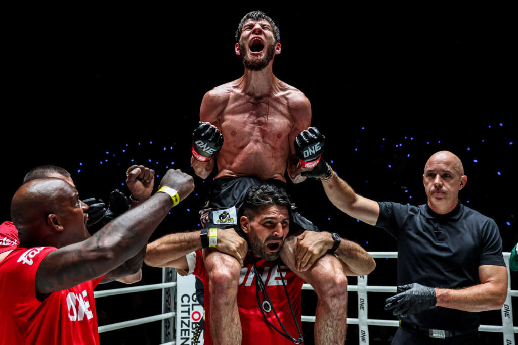 Male fighter shirtless, raised on teammates' shoulders in a boxing ring, arms raised in triumph against a dark background with blue lights.