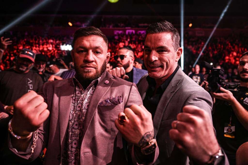 BKFC-41-Social-0199-1600x1067 - AfroCombat Two men in suits posing with fists raised at a crowded, red-lit event with photographers in the background.