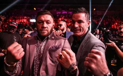 Two men in suits posing with fists raised at a crowded, red-lit event with photographers in the background.
