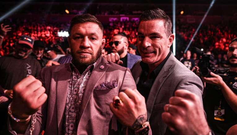 Two men in suits posing with fists raised at a crowded, red-lit event with photographers in the background.