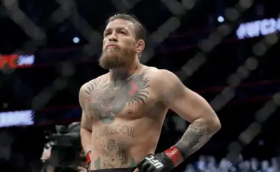 Male UFC fighter with a full beard stands in the octagon, hands on hips, wearing red UFC gloves amid a dimly lit cage. Tattoos cover his chest and arms.