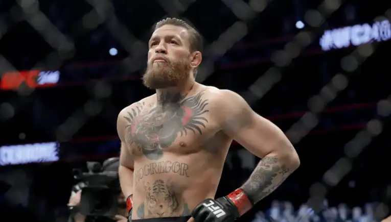 Male UFC fighter with a full beard stands in the octagon, hands on hips, wearing red UFC gloves amid a dimly lit cage. Tattoos cover his chest and arms.
