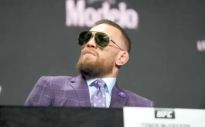Bearded man in a purple pinstripe suit and aviator sunglasses at a press conference, looking to the side with a confident pose.