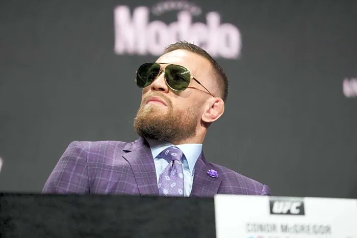 Bearded man in a purple pinstripe suit and aviator sunglasses at a press conference, looking to the side with a confident pose.