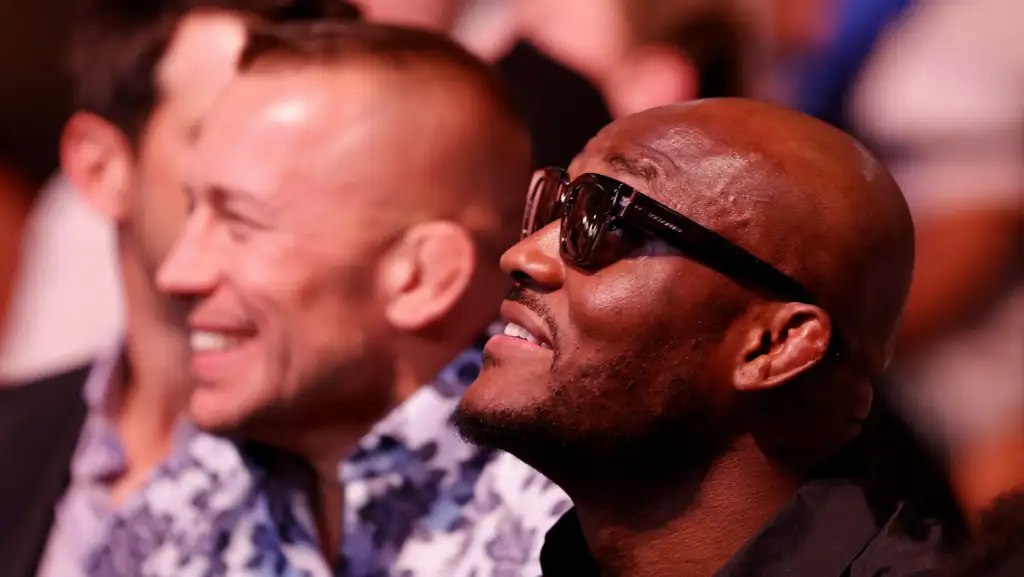 Man wearing sunglasses and a smile in the foreground at an event, with another man blurred in the background.