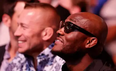 Man wearing sunglasses and a smile in the foreground at an event, with another man blurred in the background.