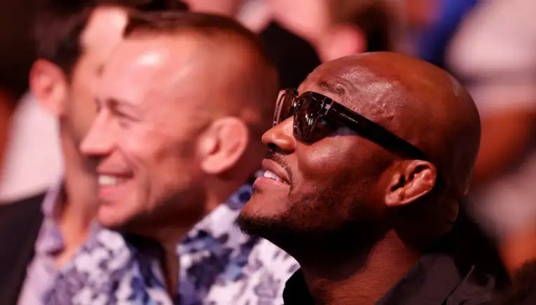 Man wearing sunglasses and a smile in the foreground at an event, with another man blurred in the background.