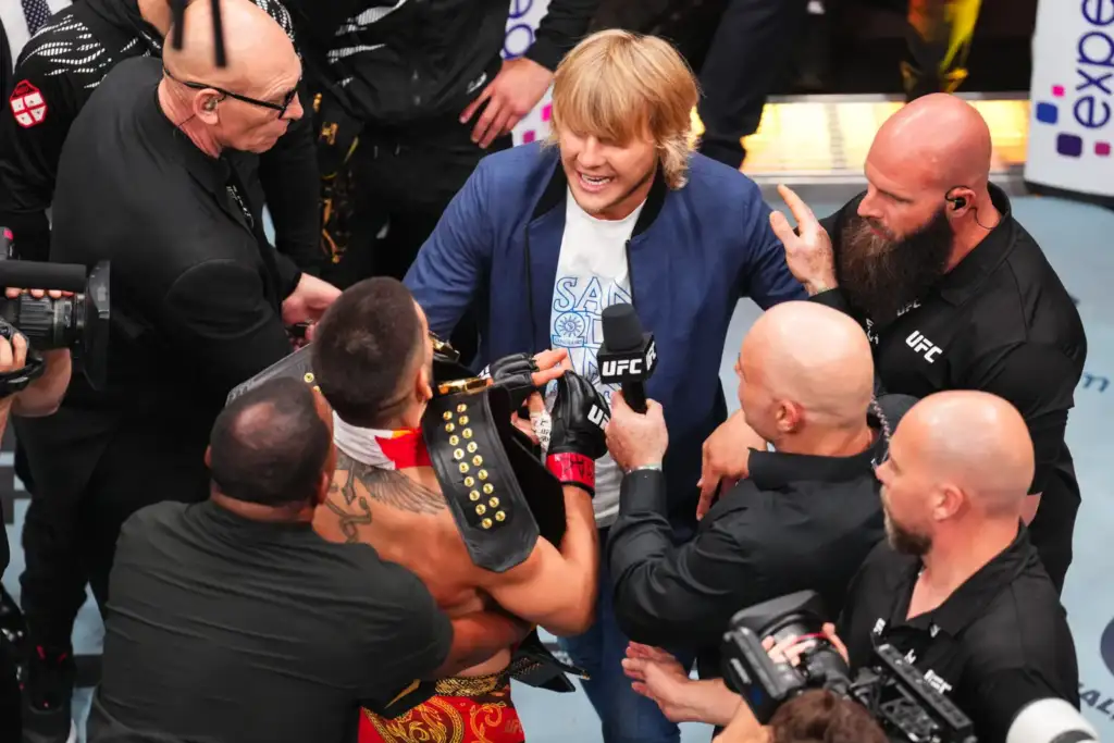 UFC fighter wearing a championship belt being interviewed by a man with a UFC microphone, surrounded by security and reporters.