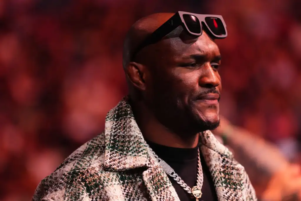Close-up of a man in a patterned coat with sunglasses on his head, against a red-toned crowd. Fixated expression, looking to the right.