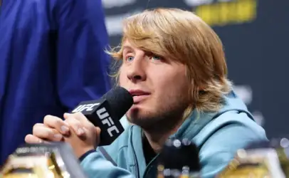 Male UFC fighter with blond hair speaking into a microphone at a press conference, wearing a light blue hoodie, with trophies visible in the foreground.