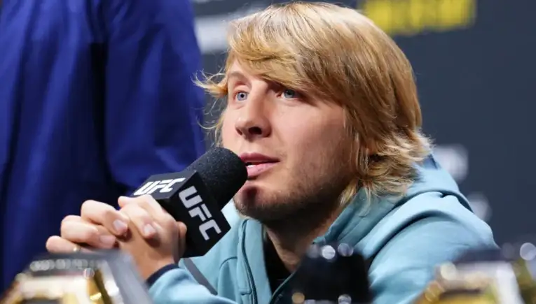 Male UFC fighter with blond hair speaking into a microphone at a press conference, wearing a light blue hoodie, with trophies visible in the foreground.