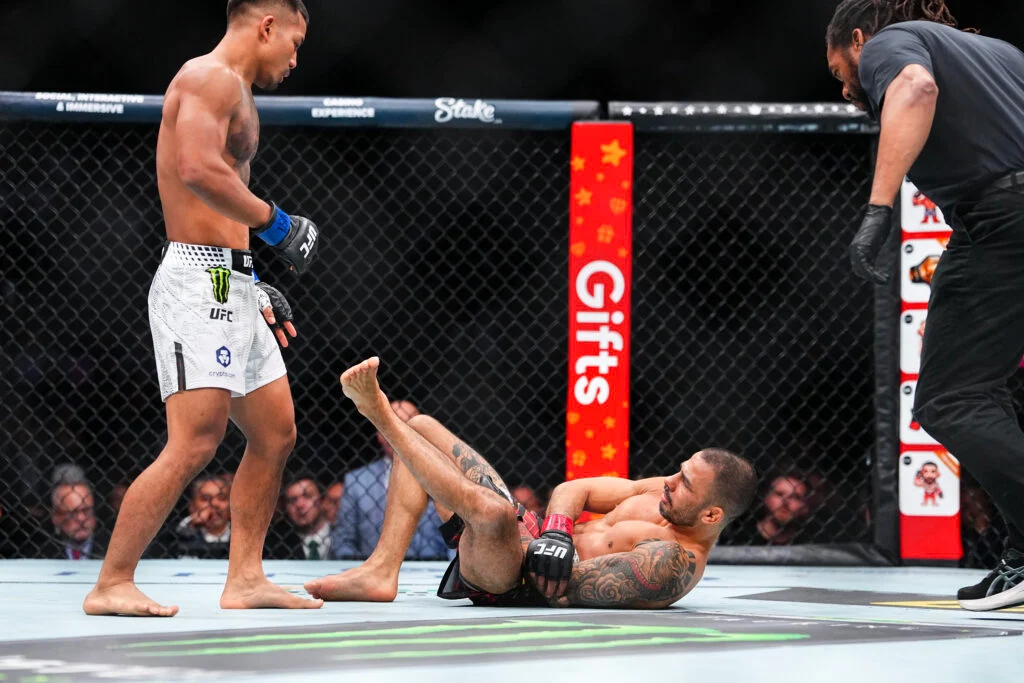 GettyImages-2250450800-1024x683 - AfroCombat UFC fighter stands over opponent on the mat inside a cage, referee approaching from the right.