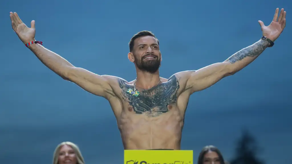 Bearded tattooed man shirtless with arms raised in triumph on a blue backdrop, yellow sign in front and spectators at the bottom left and right.