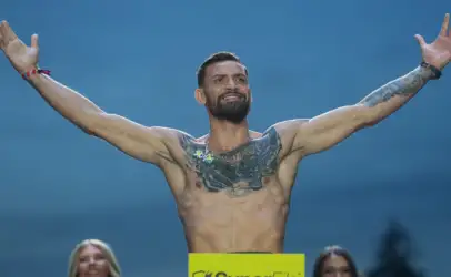 Bearded tattooed man shirtless with arms raised in triumph on a blue backdrop, yellow sign in front and spectators at the bottom left and right.