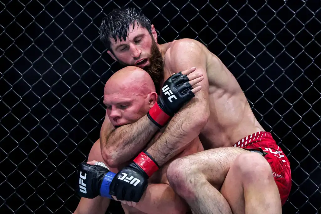 Two UFC fighters grapple in a tight clinch inside a black chain-link cage, red trunks visible in the background.