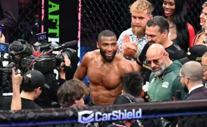 UFC fighter with a beard smiles shirtless in the ring, surrounded by cameras and teammates near a CarShield banner.