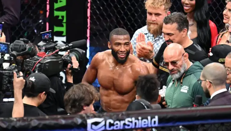 UFC fighter with a beard smiles shirtless in the ring, surrounded by cameras and teammates near a CarShield banner.