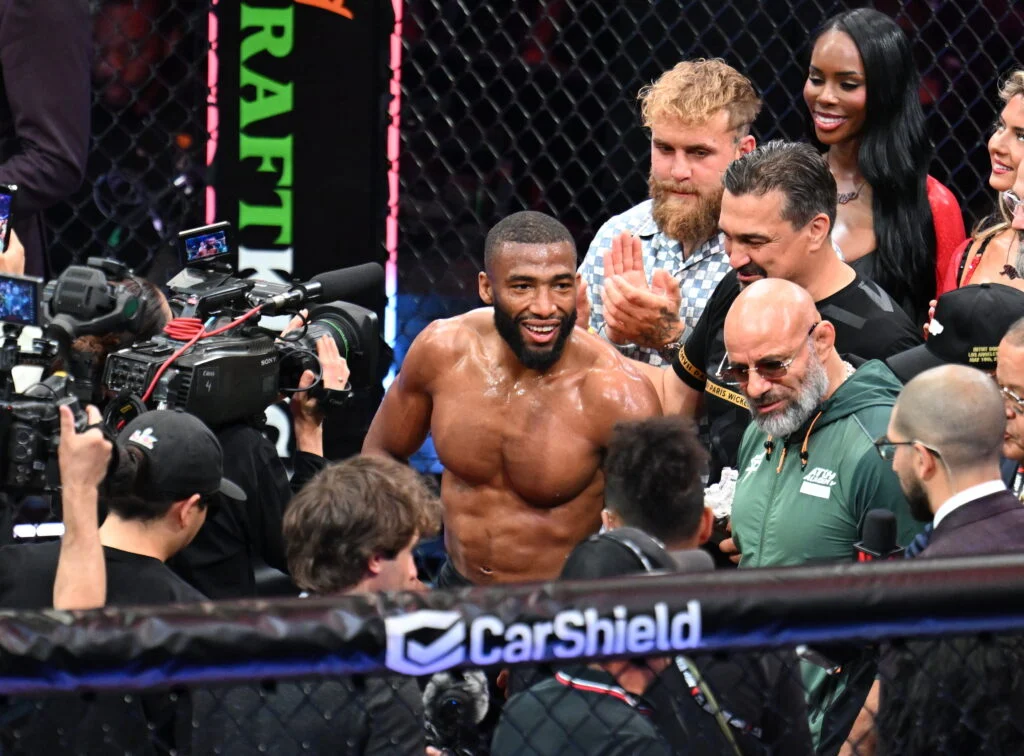 UFC fighter with a beard smiles shirtless in the ring, surrounded by cameras and teammates near a CarShield banner.