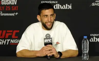 UFC fighter seated at a press conference table, smiling, holding a UFC-branded microphone; wearing a white T-shirt, with a water bottle on the table to his right.