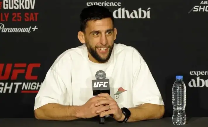UFC fighter seated at a press conference table, smiling, holding a UFC-branded microphone; wearing a white T-shirt, with a water bottle on the table to his right.