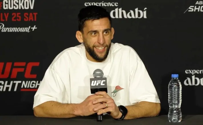 UFC fighter seated at a press conference table, smiling, holding a UFC-branded microphone; wearing a white T-shirt, with a water bottle on the table to his right.