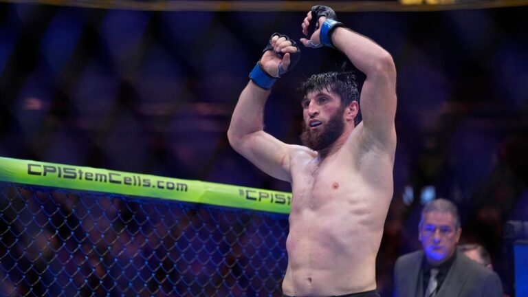 Male mixed martial arts fighter in the octagon raises his arms in victory, shirtless and sweaty, wearing blue gloves.