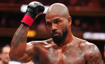 Boxer with shaved head and tattoos raises a gloved fist in the boxing ring, looking focused and intense.