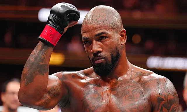Boxer with shaved head and tattoos raises a gloved fist in the boxing ring, looking focused and intense.