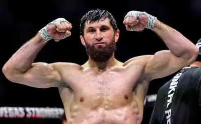 Bearded male mixed martial artist flexing his arms in the ring, sweat on his chest and fists raised in victory gesture.
