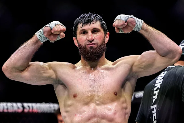 Bearded male mixed martial artist flexing his arms in the ring, sweat on his chest and fists raised in victory gesture.