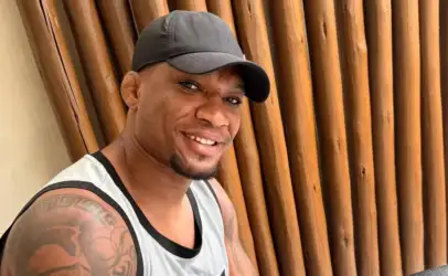 Smiling man in a gray sleeveless shirt and dark cap, standing against a wooden slat wall outdoors.