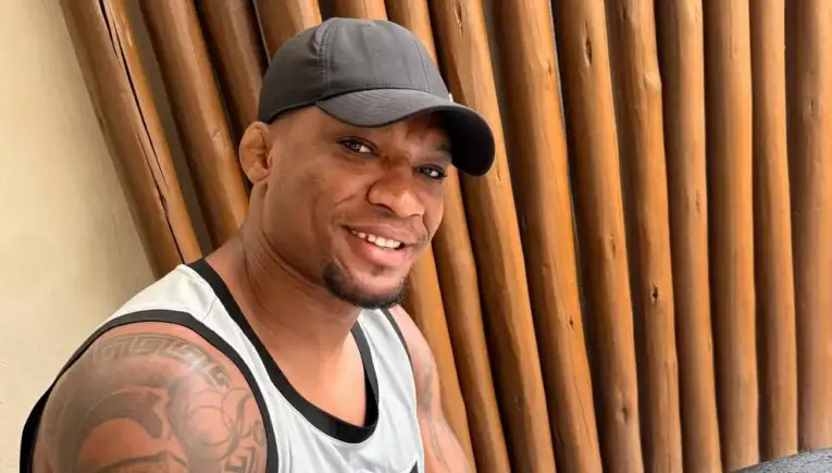 Smiling man in a gray sleeveless shirt and dark cap, standing against a wooden slat wall outdoors.