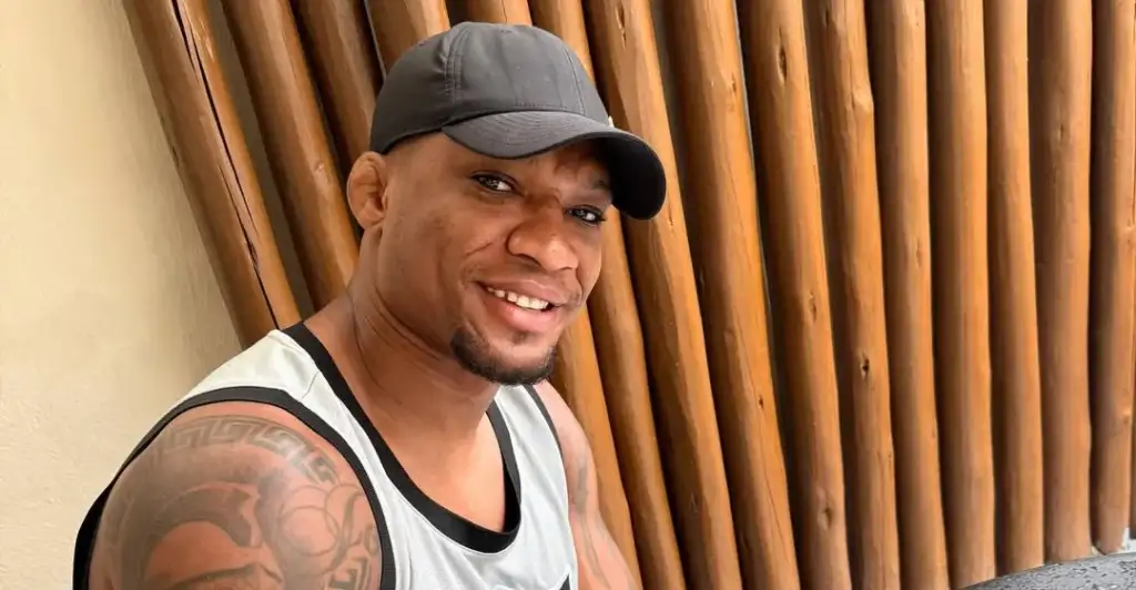 almeoda - AfroCombat Smiling man wearing a gray cap and sleeveless shirt, leaning against a vertical wooden fence.