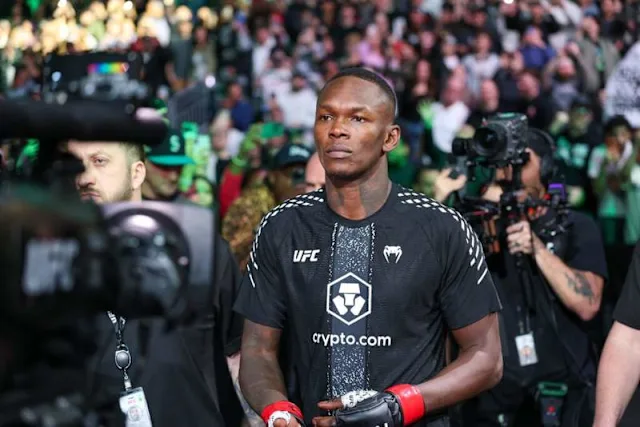 UFC fighter walks through a crowded arena, wearing a black fight shirt with the crypto.com logo, surrounded by photographers and fans.