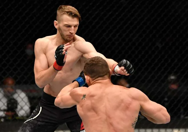 Two shirtless MMA fighters in a fenced cage; one wearing red gloves delivers a punch toward the opponent in blue gloves.