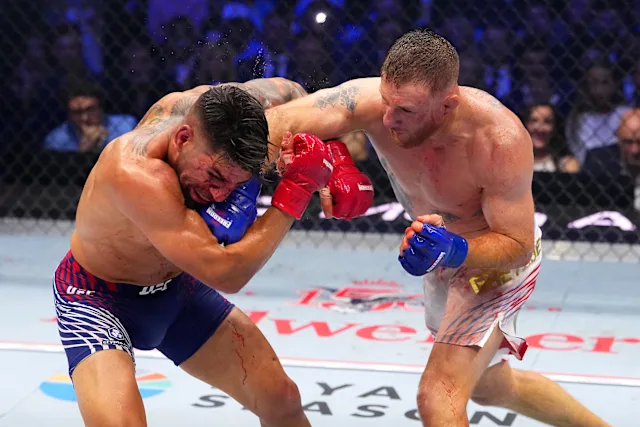 Two mixed martial artists in a UFC bout inside the cage, trading punches with blue gloves against red gloves.