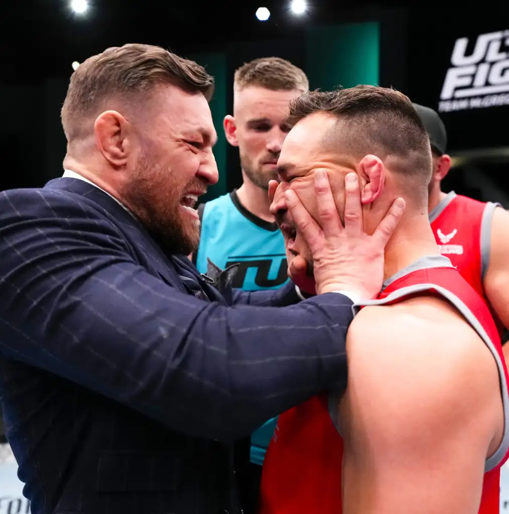 Two men share a close, comforting hug: one in a dark suit with a beard holding the other man’s head in his hands at a sports event.
