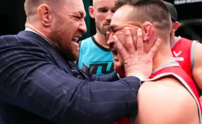 Two men share a close, comforting hug: one in a dark suit with a beard holding the other man’s head in his hands at a sports event.