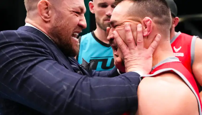 Two men share a close, comforting hug: one in a dark suit with a beard holding the other man’s head in his hands at a sports event.