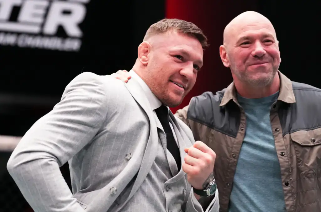 Two men posing together and smiling at the camera; the left man wears a light gray suit and black tie, the right man wears a blue t‑shirt with a brown and gray jacket.