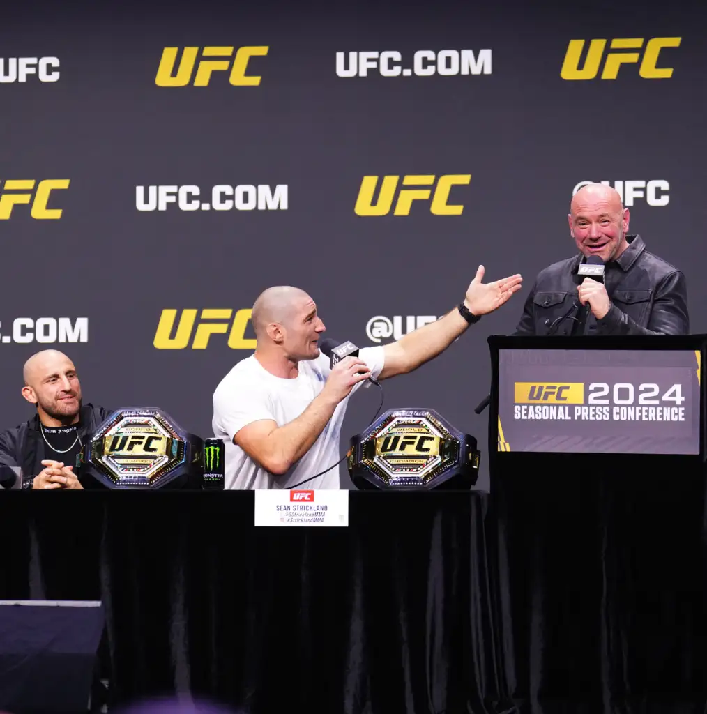 UFC press conference stage with fighters, microphones, and championship belts on the table in front of a dark backdrop with UFC logos
