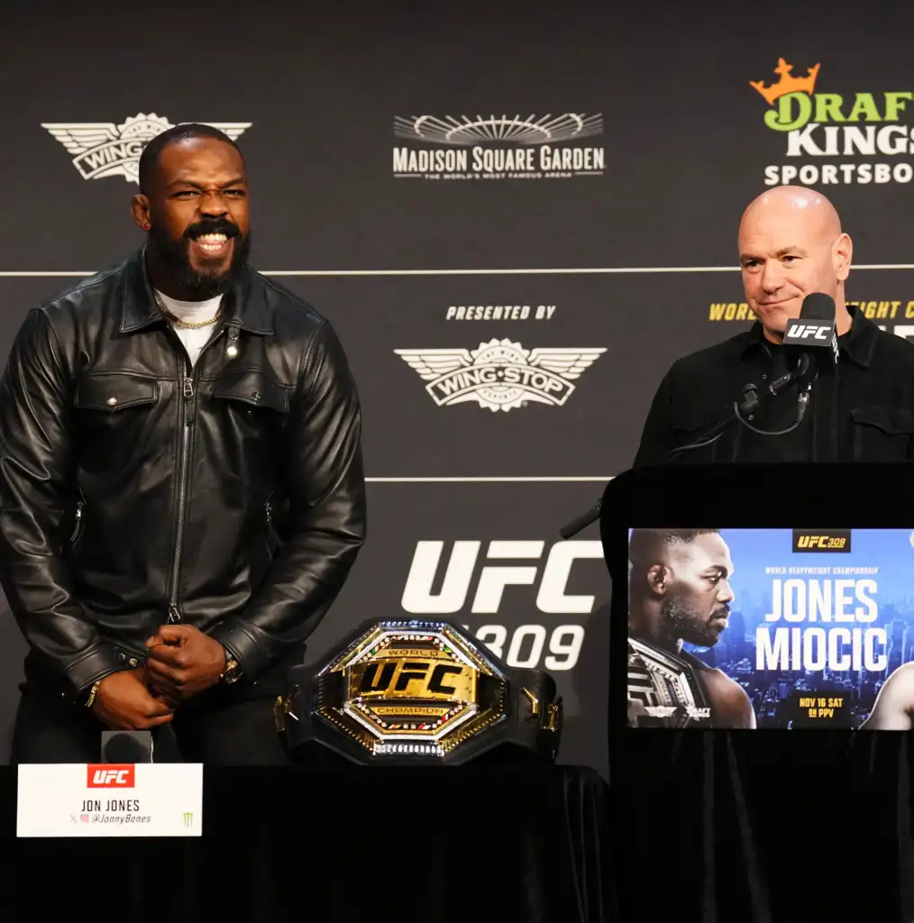 Two men at a UFC press conference with UFC logos, a championship belt on the table, and event posters in the background.