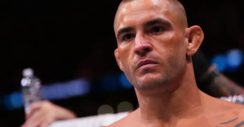 Close-up of a shirtless male fighter with short hair and a serious expression, sweat on his face, in a dimly lit arena.