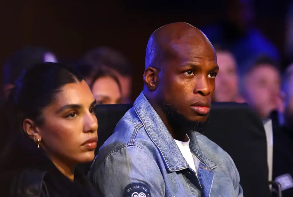 gettyimages-2241964727 - AfroCombat Man in a light denim jacket seated in a dimly lit audience, looking toward the stage; a woman with earrings sits beside him in the foreground.