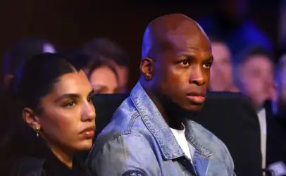 Man in a light denim jacket seated in a dimly lit audience, looking toward the stage; a woman with earrings sits beside him in the foreground.
